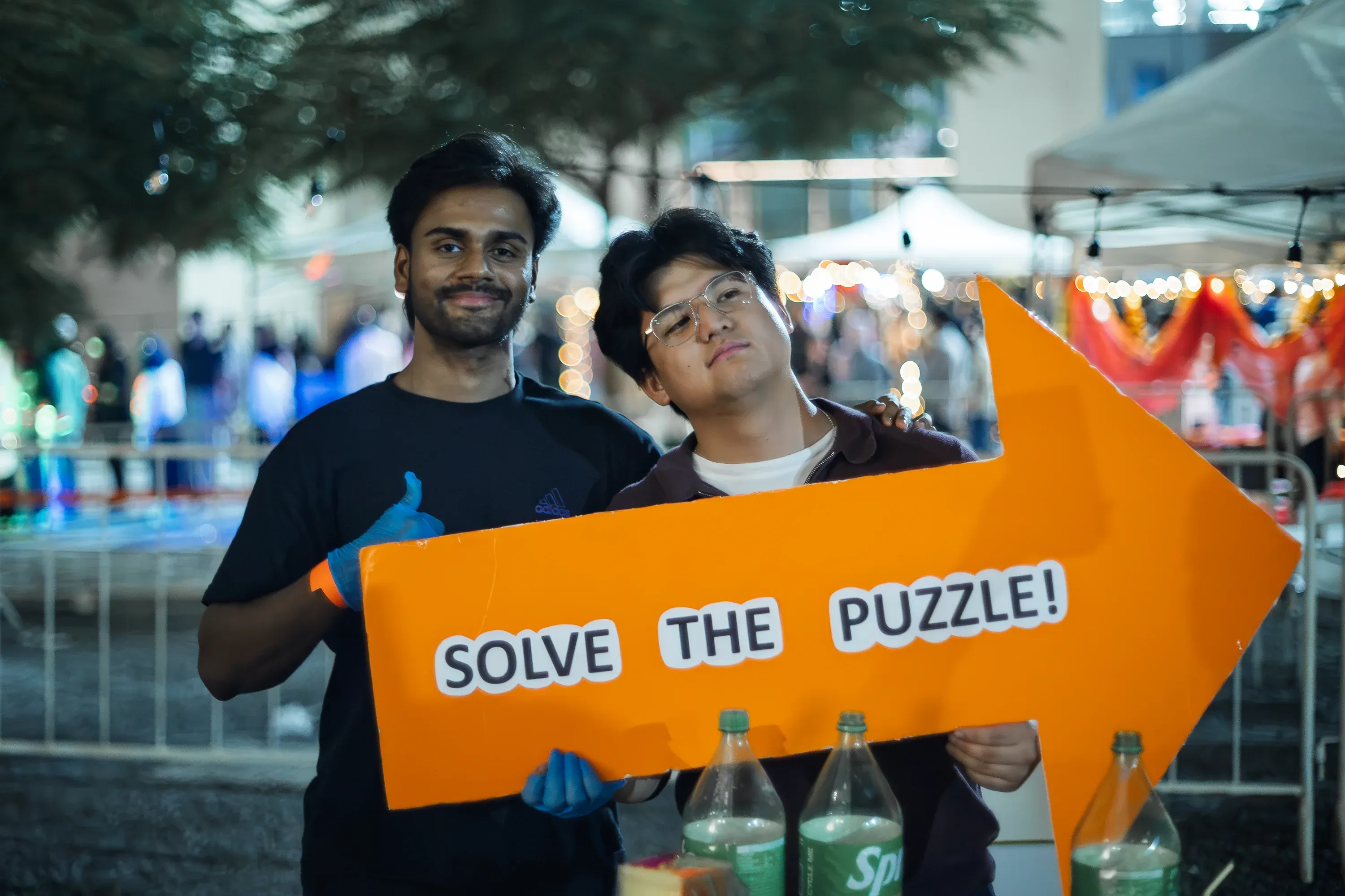 Two young men holding an orange arrow sign that says "SOLVE THE PUZZLE!" at a night event, bokeh lighting from background stalls.