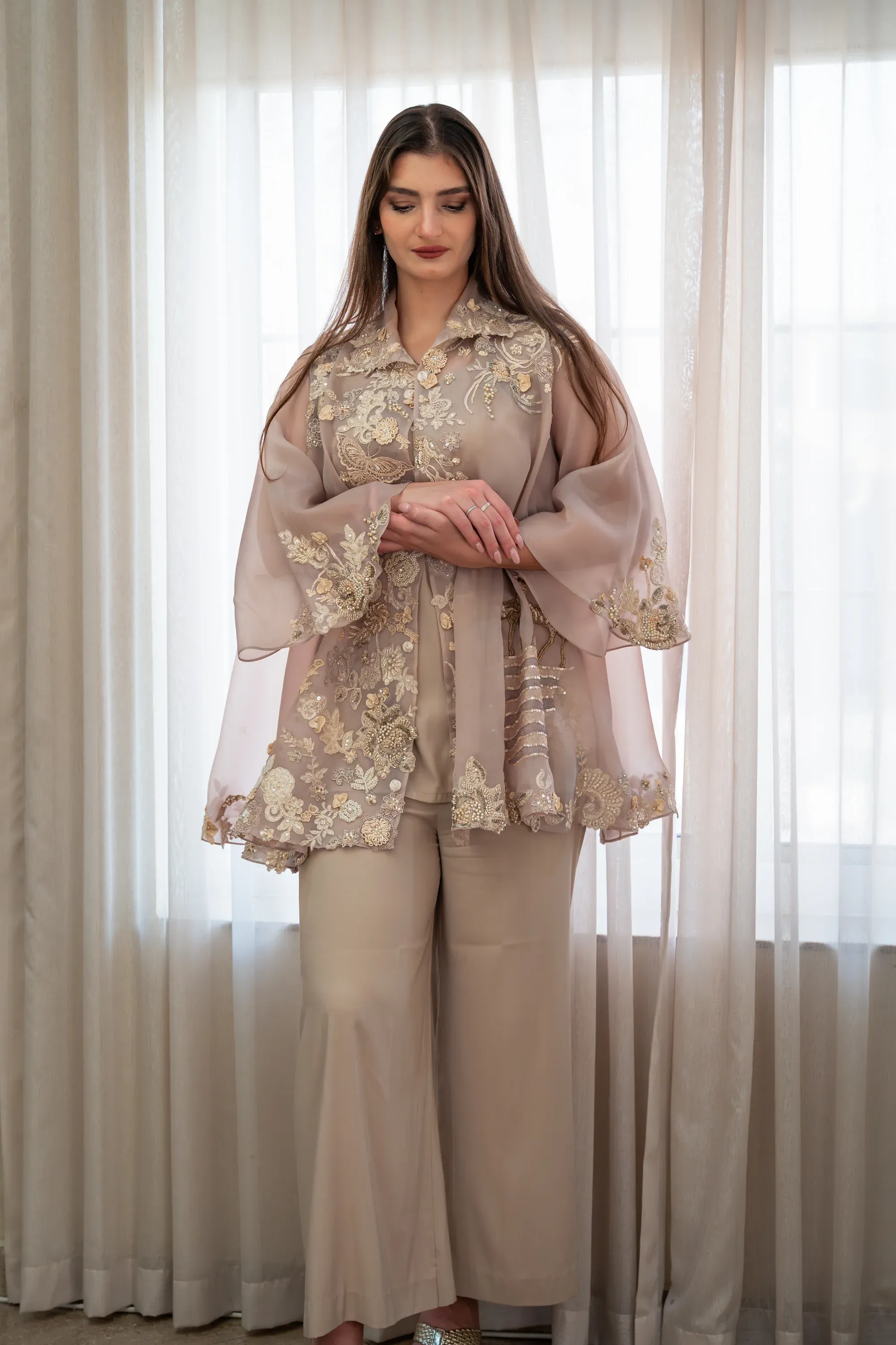 Model in an elegant embroidered beige outfit posing in front of white curtains, soft diffused window light.